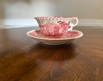 Può includere: Un set vintage di tazza e piattino da tè. La tazza e il piattino sono bianchi con un disegno di scena pastorale rosso. La tazza ha un manico e il piattino ha un bordo smerlato. Il set è posto su un tavolo di legno scuro.
