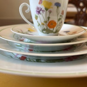 May include: A white teacup and saucer set with a floral design. The teacup features a handle and is decorated with colorful flowers and a butterfly. The saucer and plates have a green border and floral accents, creating a delicate and elegant aesthetic.