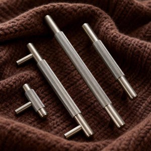 May include: A collection of four brushed nickel cabinet pulls and handles. The hardware features a ribbed design on the main body and a simple, cylindrical handle. The items are arranged on a brown knitted fabric.