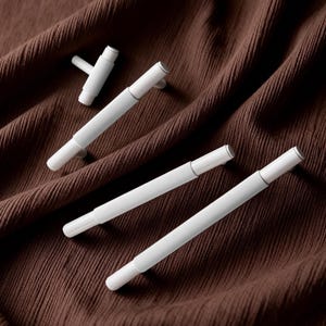 May include: Four white cabinet pulls and a knob are arranged on a brown fabric. The pulls have a cylindrical design with a ribbed texture and a small, round handle. The knob has a similar design.