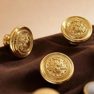 May include: Three gold-coloured cabinet knobs with a floral design are displayed. The knobs are round with an embossed pattern. They are set on a brown fabric surface, with a neutral background.