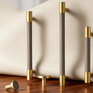 May include: A collection of cabinet hardware featuring brass and gray leather accents. The set includes several long pulls, a small knob, and a round pull. The hardware is designed for furniture and cabinetry.