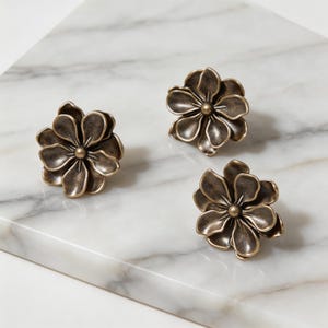 May include: Three bronze-colored flower-shaped cabinet knobs. Each knob features detailed petals and a central, rounded accent. The knobs are arranged on a white and gray marble surface, offering a decorative hardware option.