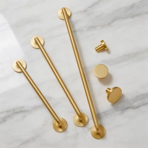 May include: A collection of gold-colored bathroom hardware, including three towel bars of varying lengths and three knobs. The bars have circular mounting plates and are straight. The knobs are round and cylindrical. The hardware is displayed on a white marble surface.
