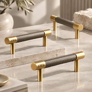 Puede incluir: Tres tiradores de armario con un marco cilíndrico de metal dorado cepillado y una sección central envuelta en cuero gris oscuro. Cada extremo de la barra de metal dorado está grabado con las letras JML. El herraje tiene dos soportes verticales.