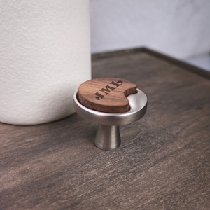 May include: A cabinet knob featuring a brushed silver metal base and a circular dark brown wood inlay. The wood inlay is engraved with the letters JML in a serif font. The knob has a tapered stem and a flat, circular top surface.