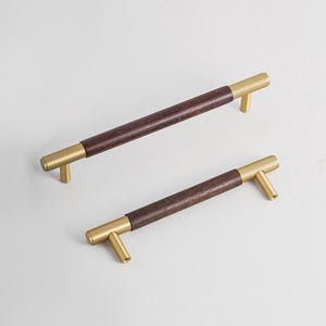 Peut inclure: Deux poign&eacute;es de meubles avec un centre en bois brun fonc&eacute; et des extr&eacute;mit&eacute;s en m&eacute;tal dor&eacute;. Les poign&eacute;es sont rectangulaires avec un design en forme de T &agrave; chaque extr&eacute;mit&eacute;. Fond blanc.