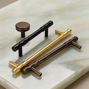 Könnte beinhalten: Eine Sammlung von Schrankbeschlägen in verschiedenen Ausführungen, darunter Schwarz, Gold und Bronze. Die Beschläge weisen geometrische Facettendesigns auf und umfassen Knöpfe und Stangengriffe. Die Artikel werden auf einer hellen, marmorähnlichen Oberfläche präsentiert.
