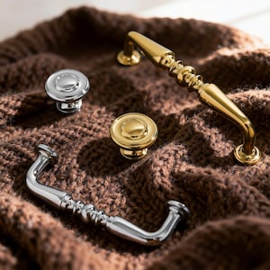 Puede incluir: Una variedad de herrajes para armarios, que incluyen tiradores y pomos plateados y dorados. Los artículos se presentan sobre una superficie marrón texturizada, destacando los diferentes estilos y acabados disponibles.