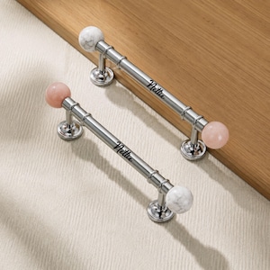 May include: Two silver-toned metal cabinet pulls with cylindrical bars and spherical ends. One pull features white marble-patterned spheres, while the other has light pink spheres. Each bar is engraved with the name Nettie in black script.