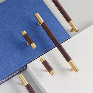 Peut inclure: Ensemble de poign&eacute;es et de tirettes de meubles en bois avec des d&eacute;tails en laiton. Les poign&eacute;es ont un centre en bois rectangulaire brun fonc&eacute; avec des embouts et des fixations en laiton. Le mot &laquo; D&eacute;coration &raquo; est visible sur un livre bleu.