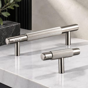 May include: Two metallic cabinet pulls in a gunmetal finish, featuring a central ribbed texture and smooth ends. Each pull is engraved with the letters JML on the side. The hardware consists of a long bar handle and a smaller T-shaped knob.