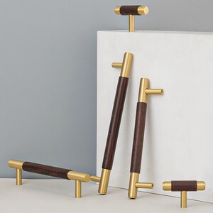 Peut inclure: Ensemble de quincaillerie de meubles moderne, combinant du bois brun fonc&eacute; et du m&eacute;tal dor&eacute; bross&eacute;. L'ensemble comprend des poign&eacute;es et des boutons de diff&eacute;rentes tailles, mettant en valeur un design minimaliste. Les poign&eacute;es et les boutons sont en cm.