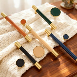 May include: A collection of cabinet hardware, including handles and knobs. The handles feature gold-toned metal ends with leather or painted wood centers in brown, black, green, blue, and natural wood tones. The knobs are round and match the handle colors.