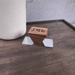 May include: A trapezoidal wooden handle with the letters JML engraved on the top surface. The handle is attached to a rectangular, reflective silver-colored metal base plate. The wood features a natural brown grain pattern and smooth finish.