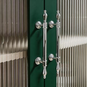 Puede incluir: Dos manijas de puerta de cromo pulido con un diseño clásico de remate, montadas en un marco de puerta verde oscuro. La puerta cuenta con paneles de vidrio estriado vertical a ambos lados, creando una apariencia texturizada y semitransparente.