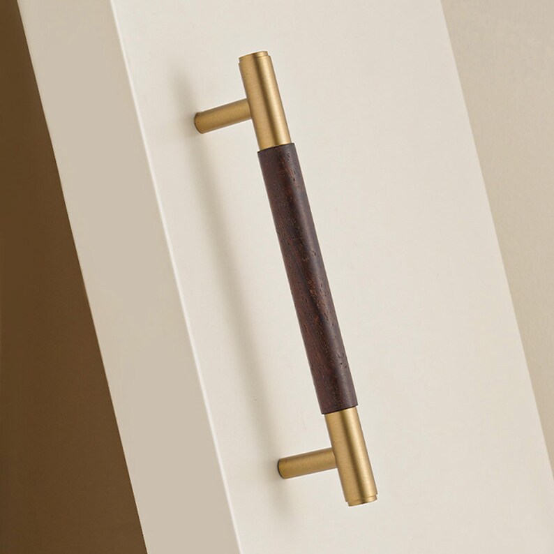 Peut inclure: Une poign&eacute;e de meuble avec un centre en bois brun fonc&eacute; et des extr&eacute;mit&eacute;s de couleur laiton. La poign&eacute;e est mont&eacute;e sur une surface blanche, mettant en valeur son design. Id&eacute;ale pour tiroirs et armoires.