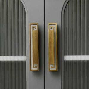 Puede incluir: Primer plano de un par de puertas de armario con tiradores rectangulares de color latón. Los tiradores tienen un diseño de clave griega con un borde blanco. Las puertas son grises con paneles de vidrio acanalado vertical.