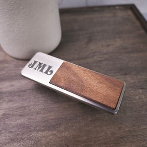 May include: A rectangular metal handle featuring a brushed silver finish and a dark brown wood inlay on one side. The metal surface is engraved with the letters JML in a stylized font. The object has a sleek, elongated shape with rounded corners.