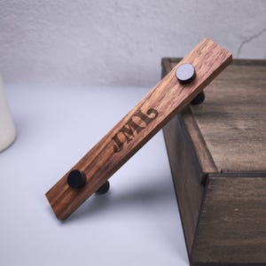 May include: A rectangular wooden drawer pull featuring a natural wood grain finish and two matte black circular metal knobs. The wood surface is engraved with the letters JML in a serif font. The hardware is positioned at an angle.