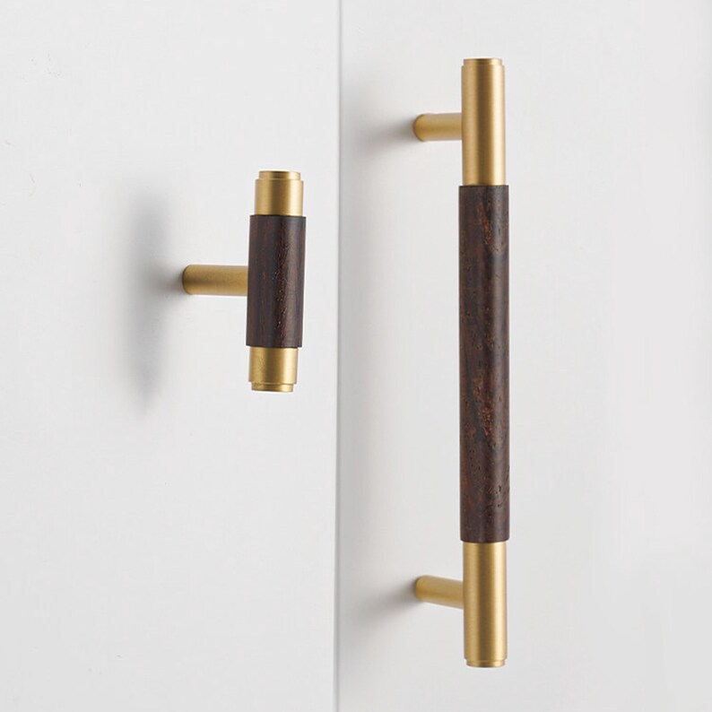 Peut inclure: Ensemble de quincaillerie d'armoire avec un centre en bois brun fonc&eacute; et des accents m&eacute;talliques dor&eacute;s. L'ensemble comprend un petit bouton et une longue poign&eacute;e, tous deux mont&eacute;s sur une surface blanche. La quincaillerie est de style moderne.