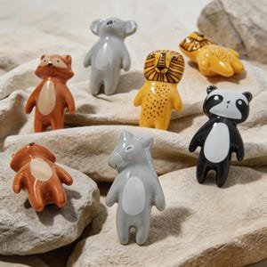 Könnte beinhalten: Eine Sammlung von Keramik-Tierfiguren, darunter ein Fuchs, ein Koala, ein Löwe, ein Panda und ein Nashorn. Die Figuren sind in verschiedenen Farben wie Orange, Grau, Gelb und Schwarz auf einer hellen, strukturierten Oberfläche angeordnet.