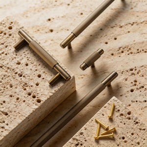 May include: Close-up of several antique brass cabinet pulls and knobs with a ridged design. The hardware is arranged on a light beige, textured surface, with a few small brass screws scattered nearby. The pulls have a T-bar design.