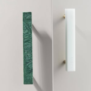 May include: Two rectangular cabinet pulls mounted on a light gray surface. One pull is dark green with a marbled texture, and the other is solid white. Both handles feature the word MACK embossed vertically along the front surface of the hardware.