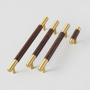 Peut inclure: Ensemble de quatre poign&eacute;es de meubles. Elles pr&eacute;sentent un centre en bois brun fonc&eacute; avec des extr&eacute;mit&eacute;s et des pieds en m&eacute;tal dor&eacute;. Les poign&eacute;es varient en taille, la plus longue mesurant environ 30 cm de long.