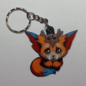 Op de afbeelding: Een kleurrijke sleutelhanger met een cartoonachtig vosachtig wezen met oranje, rode en blauwe accenten. De sleutelhanger heeft een zilveren ring en ketting. Het wezen heeft een verdrietige uitdrukking en draagt een hoofddeksel.