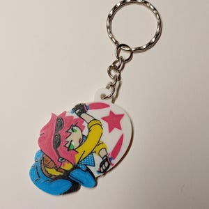 Puede incluir: Un llavero de acrílico blanco con un personaje de dibujos animados de cabello rosa brillante, que viste una camisa amarilla y pantalones azules, sosteniendo dos cuchillos rosas. El diseño incluye una estrella rosa y una anilla de metal.