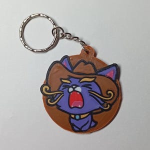 May include: A novelty keyring featuring a cartoon cat wearing a cowboy hat. The cat is purple with a wide open red mouth, gold accents, and a blue collar. The keyring has a silver ring and chain.