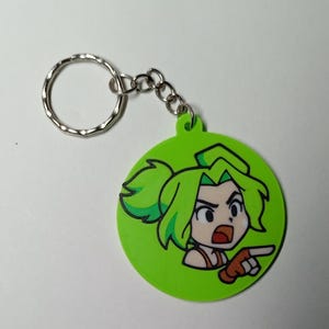 May include: A green circular keyring featuring a cartoon character with green hair and a pointing gesture. The character is set against a green background, with a silver key ring and chain attached.