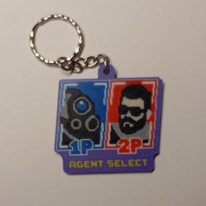 Könnte beinhalten: Ein lila Schlüsselanhänger mit einem pixeligen Design, das zwei Charaktere in blauen und roten Quadraten zeigt. Der Text "1P" und "2P" ist sichtbar, zusammen mit "AGENT SELECT". Der Schlüsselanhänger hat einen silbernen Ring.
