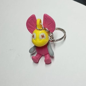 May include: A small, colorful keychain featuring a cartoon character with a yellow head, pink ears and body, and gray wings. The character has cross-shaped eyes and is attached to a silver keyring. The keychain is made of plastic.
