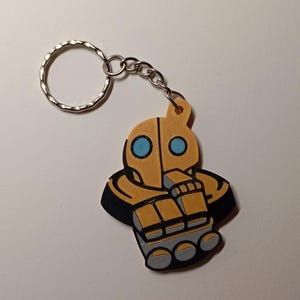 Könnte beinhalten: Ein Schlüsselanhänger mit einer stilisierten, cartoonartigen Figur in Gelb-, Schwarz- und Grautönen. Die Figur hat blaue Augen und ist an einem kreisförmigen Schlüsselring befestigt. Das Design erinnert an einen Roboter oder eine futuristische Figur.