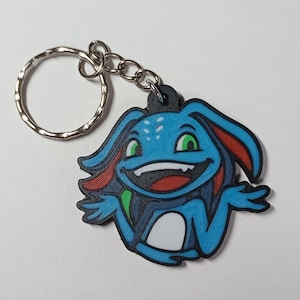 May include: A novelty keychain featuring a cartoon character in blue, red, and black, with a silver keyring. The character has a smiling face with green eyes and a white belly. A fun accessory.