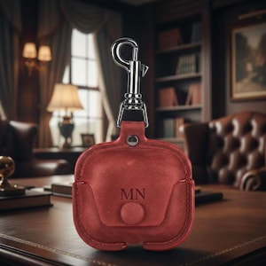 May include: A red leather AirPod case with a silver carabiner clip. The case has a flap closure and the initials "MN" are embossed on the front. The case is sitting on a dark wooden desk.