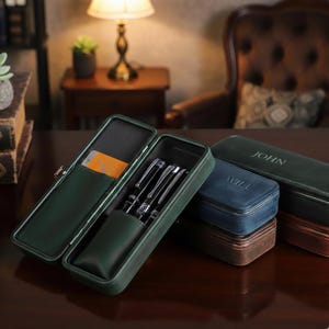 Op de afbeelding: Een open, donkergroene leren pennenetui met drie zilveren pennen erin. Een creditcard is zichtbaar in een zijvak. Andere gesloten etuis in blauw, groen en bruin zijn ook aanwezig, met de namen "JOHN" en "WILL" erin geëmbosseerd.