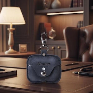 May include: A navy blue leather AirPod case with a silver clasp and button closure. The name "Hunter" is embossed on the front. The case is designed to protect and carry wireless earbuds. The case is shown on a wooden desk.