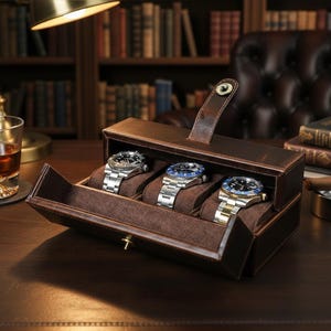 May include: A brown leather watch case with three silver and black watches. The case is open, revealing the watches on brown velvet cushions. A gold-toned lamp and a glass of amber liquid are in the background.