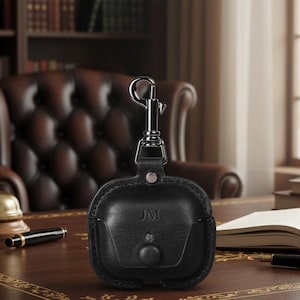 May include: Black leather AirPod case with silver-tone clasp and the initials "JM" embossed on the front. The case has a snap closure and is shown on a dark wood desk with a pen and open book.