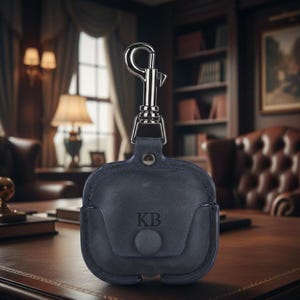 May include: Navy blue leather AirPod case with a silver-tone carabiner clip. The case features the initials "KB" embossed on the front and a snap closure. The case is designed to protect and carry wireless earbuds. The overall aesthetic is classic and sophisticated.