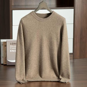 Può includere: Un maglione beige girocollo a maniche lunghe è appeso a una gruccia di legno. Il maglione ha scollo, polsini e orlo a coste. Lo sfondo comprende una mensola di legno e dei libri.