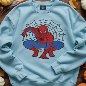 Peut inclure: Un sweat-shirt bleu clair avec un motif de Spider-Man accroupi devant une toile d'araignée noire. Le super-héros est représenté dans son costume rouge et bleu classique. Le sweat-shirt a un col rond et des manches longues.