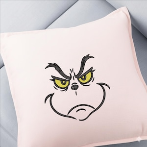May include: A light pink square cushion featuring an embroidered Grinch face. The Grinch's face is outlined in black with yellow eyes and a grumpy expression. The cushion is on a light blue surface.