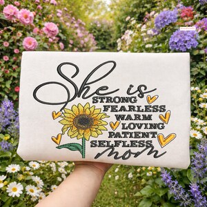 May include: Cream-colored rectangular pillow with the embroidered text "She is strong, fearless, warm, loving, patient, selfless mom." A yellow sunflower and heart accents are also embroidered on the pillow.
