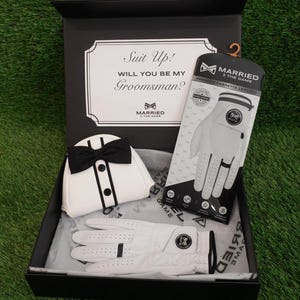 May include: A black gift box containing a white golf glove with black trim, a matching white golf glove in its retail packaging, and a white golf club head cover featuring a black bow tie and buttons. The box lid reads Suit Up! WILL YOU BE MY Groomsman?