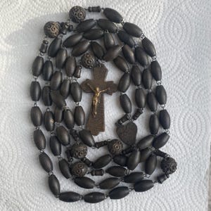 May include: A dark brown rosary with oval wooden beads and decorative metal accents. The rosary features a cross with a gold-colored Jesus figure and a heart-shaped pendant. The beads are strung together with metal links.