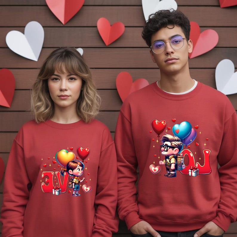 May include: Two people wearing matching red sweatshirts. Each sweatshirt features a cartoon illustration of a couple with heart-shaped balloons and the word "LOVE" in a stylized font. The background includes heart-shaped decorations.